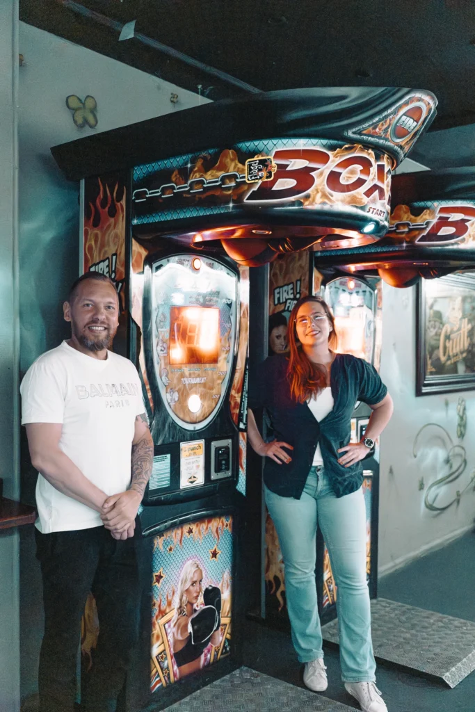 Lasse står ved siden af en boksemaskine på Old Irish Pub i en hvid t-shirt og ser glad ud, da han har hjulpet dem med at tjene penge